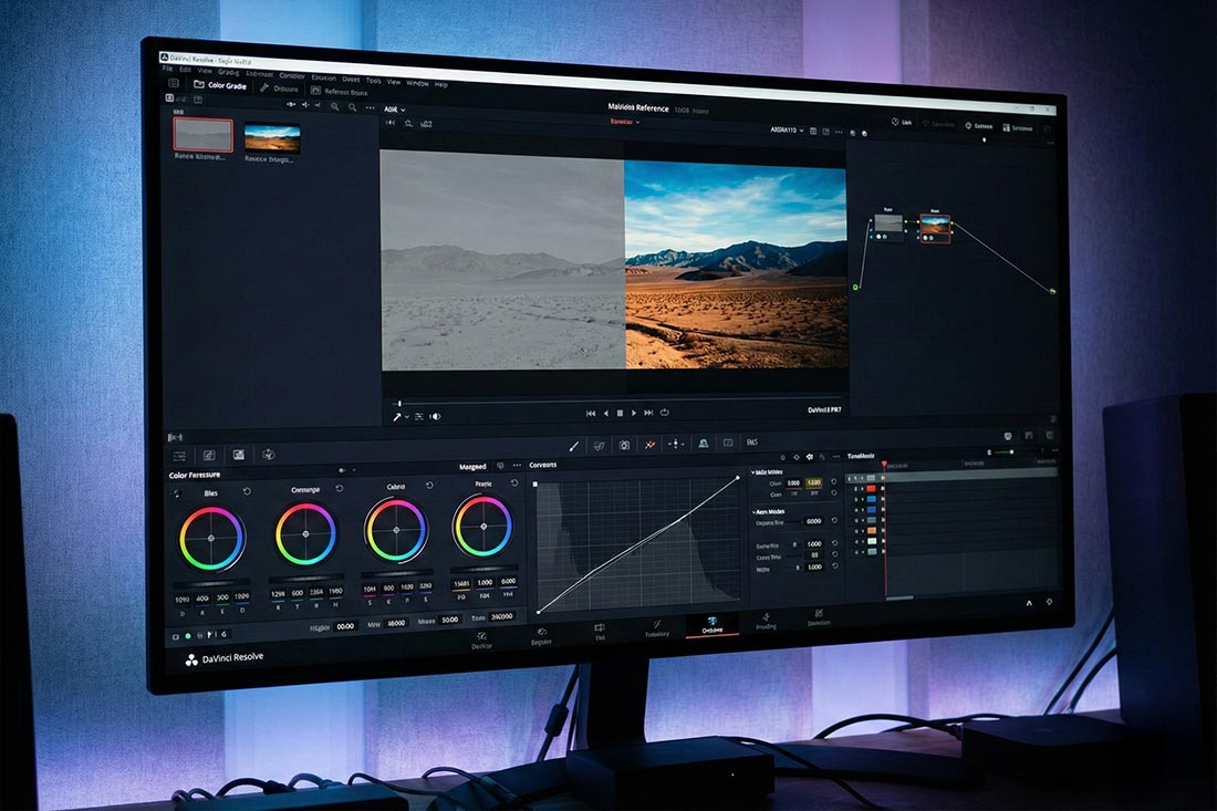 Realistyczne zdjęcie monitora komputerowego w studiu wideo. Na ekranie widać interfejs programu do koloryzacji (cyfrowe koła kolor&oacute;w i krzywe) oraz podgląd wideo z wyraźnym podziałem: lewa strona to surowy, szary obraz, a prawa to nasycony, gotowy film. Jednoznacznie wskazuje to na proces cyfrowej obr&oacute;bki obrazu na komputerze.